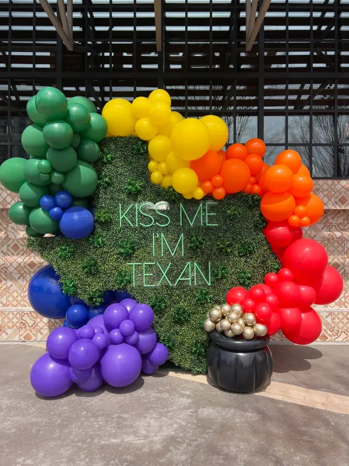 a heart shaped sign with balloons in the shape of a rainbow and the words kiss me i'm texan
