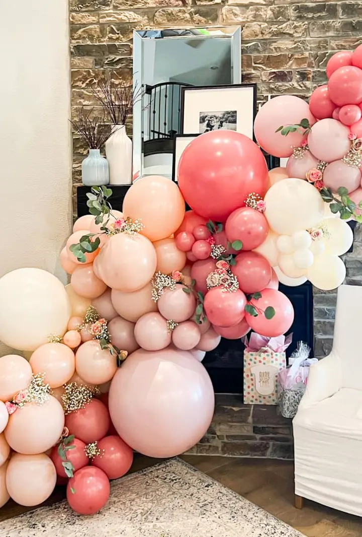 A vibrant and whimsical display of various sized balloons in shades of pink, peach, and white, arranged in a cluster against a rustic brick wall backdrop, creating a visually striking and festive scene.