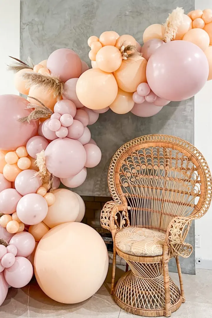 A vibrant arrangement of pink, peach, and white balloons adorns the foreground, complemented by a wicker chair in the background, creating a visually striking and whimsical scene.