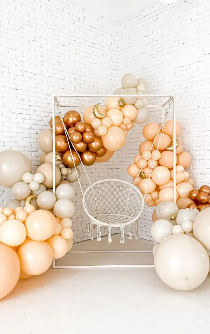 A wire frame chair surrounded by an assortment of balloons in various shades of beige, cream, and gold, set against a plain white background.