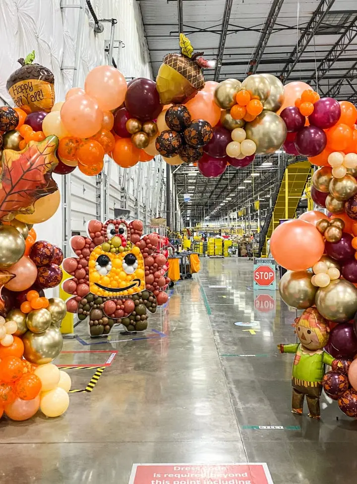 A vibrant and whimsical display of colorful balloons, autumn-themed decorations, and a smiling face sculpture, all set against a large industrial warehouse-like space.