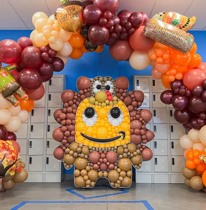 A vibrant and whimsical balloon arch made of various fruits and vegetables, including grapes, oranges, and pumpkins, stands in front of a row of lockers in what appears to be a school or office setting.