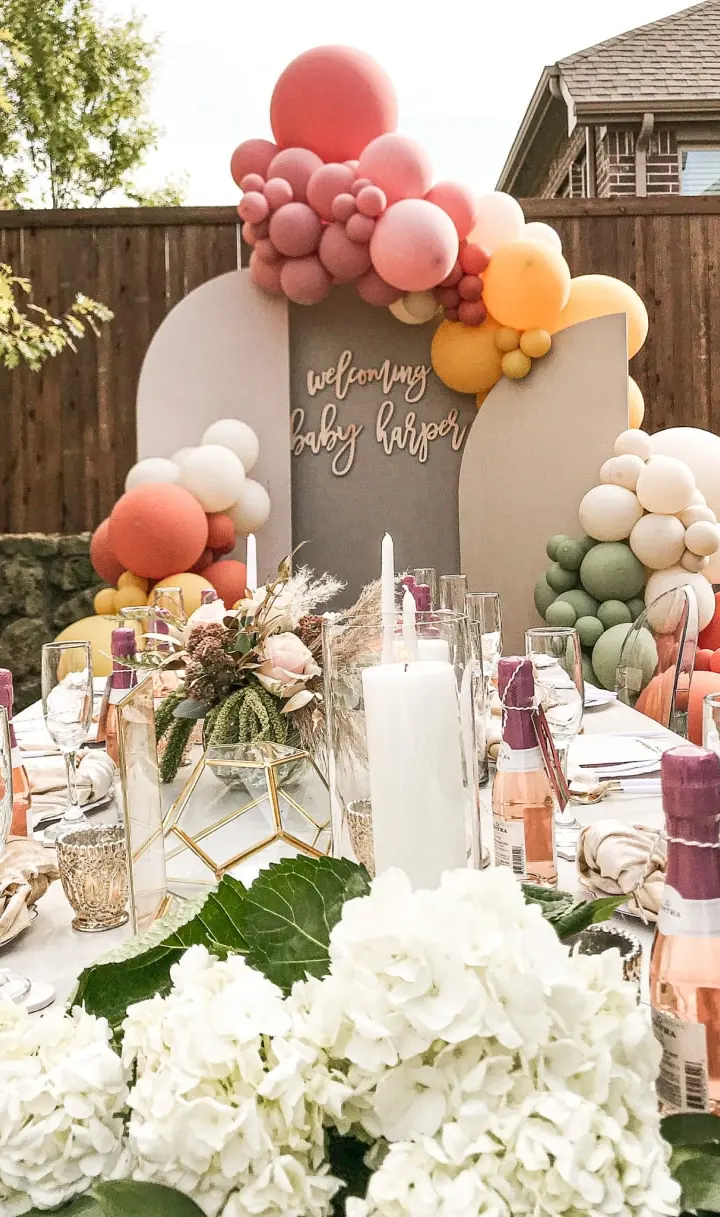 A beautifully decorated outdoor table setting with an array of colorful balloons, floral arrangements, and various decorative elements, all set against a backdrop of a wooden fence and lush greenery.