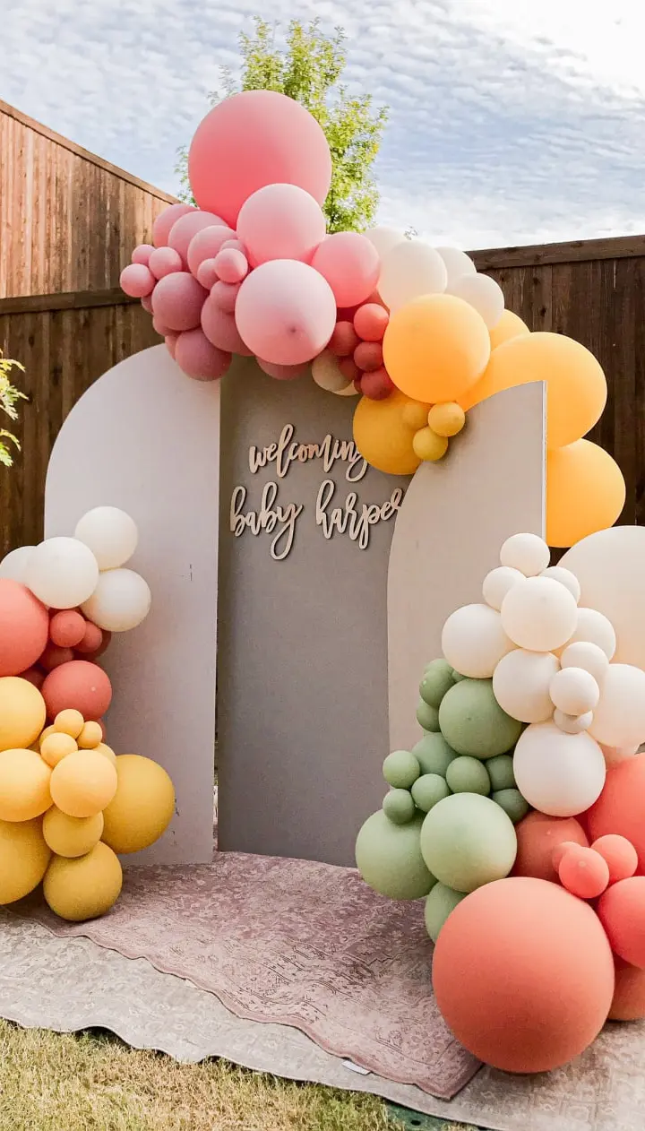 A colorful and whimsical balloon arch adorns the entrance to a baby shower event, with a variety of pastel-colored balloons of different sizes creating a vibrant and festive display against a wooden fence backdrop.