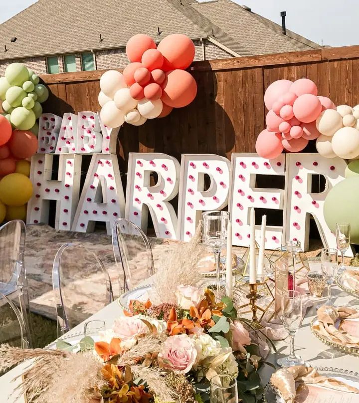 A festive outdoor setting with a large "HARPER" sign made of letters, surrounded by colorful balloons in various shades of pink, red, and white, and a table decorated with floral arrangements and place settings.
