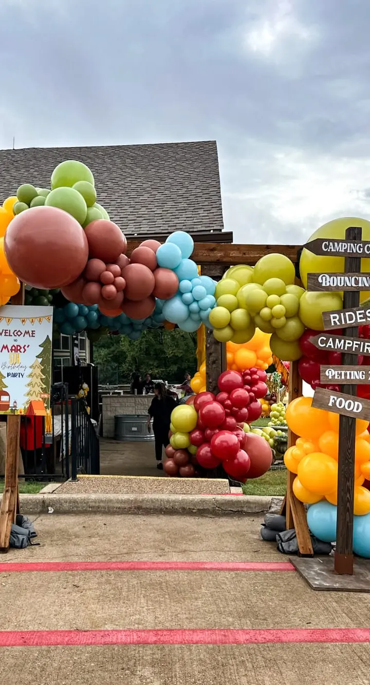 A colorful display of large inflatable balloons in various shapes and sizes, including fruits and other objects, stands in the foreground, while a wooden building with signage for a camping and recreational vehicle business can be seen in the background.