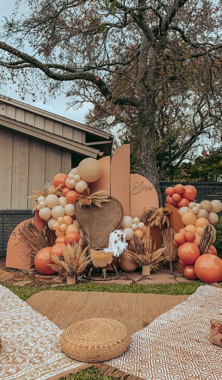 A vibrant autumn display featuring an assortment of pumpkins, gourds, and other seasonal decorations arranged in the foreground, set against a backdrop of a wooden building and a lush, tree-lined landscape.