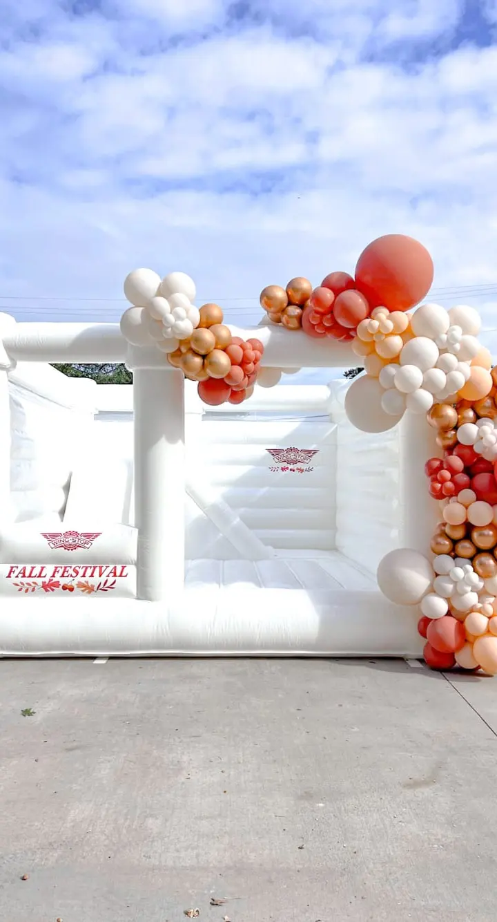 A large white inflatable structure with colorful balloon clusters decorating the exterior, set against a blue sky with scattered clouds in an outdoor setting.