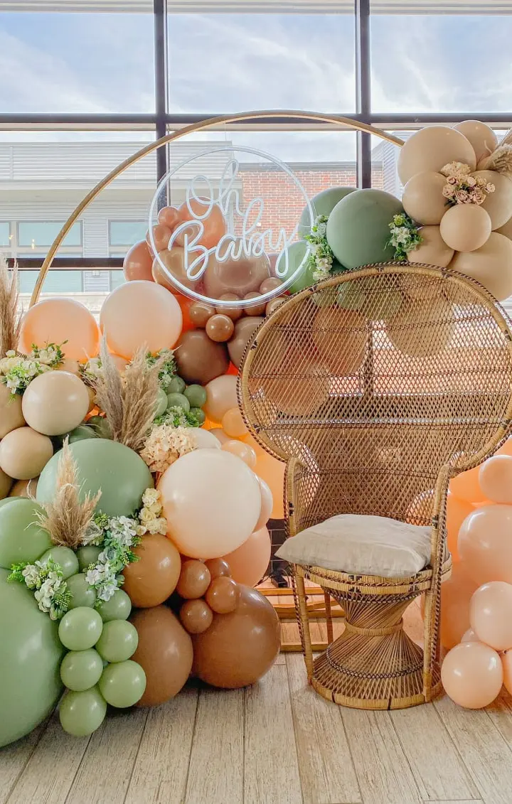 A vibrant and whimsical display of pastel-colored balloons, greenery, and a wicker chair set against a large window, creating a warm and inviting atmosphere.