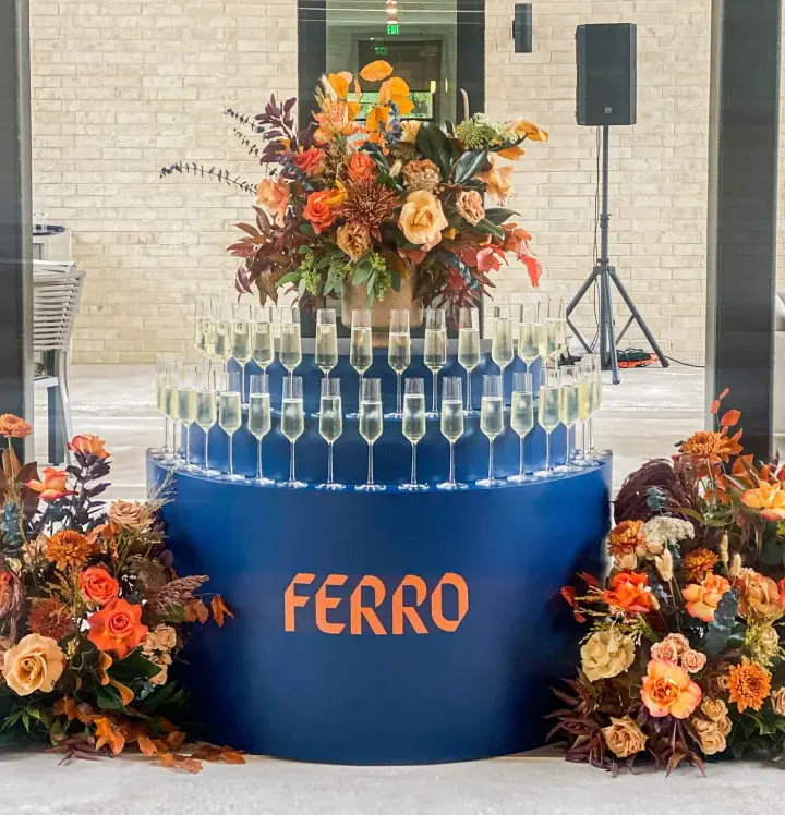 A large, ornate floral arrangement sits atop a blue platform with the word "FERRO" prominently displayed, surrounded by a display of wine glasses in a modern, industrial-style setting.