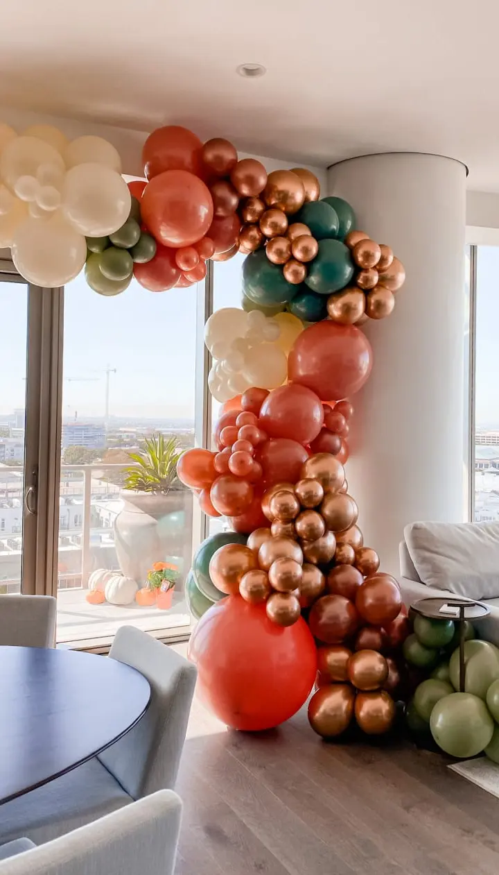 A colorful and whimsical balloon arch made of various shades of orange, red, and green balloons dominates the foreground, while a panoramic view of a city skyline can be seen through the large windows in the background.