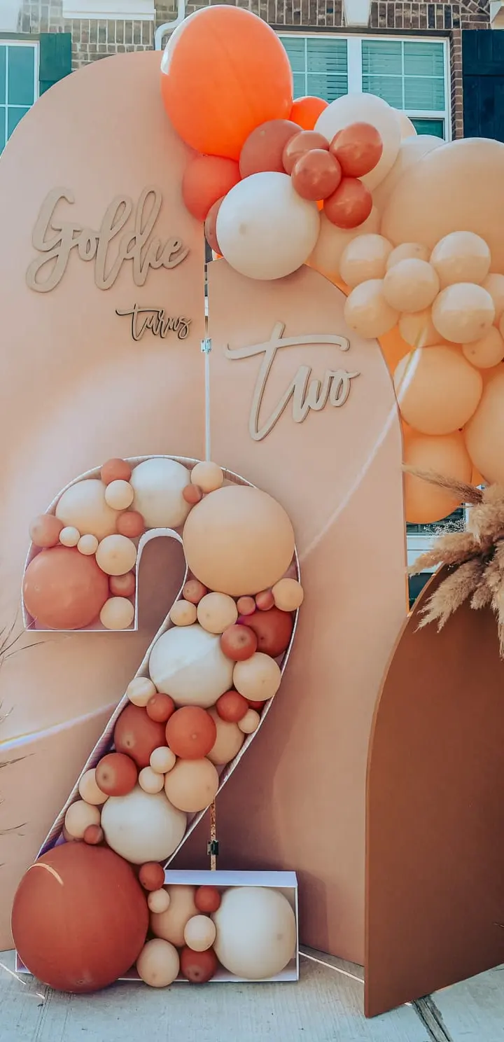The image features a large, decorative display with the number "2" prominently displayed, surrounded by various colorful balloons, pumpkins, and other autumn-themed elements against a backdrop of a brick building.