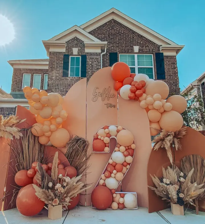 A large, colorful display of autumn-themed decorations, including balloons, pumpkins, and dried foliage, stands in the foreground, with a brick house visible in the background.