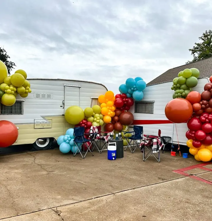 A colorful display of balloons in various shapes and sizes, including round, oblong, and elongated forms, surrounds a vintage-style camper trailer in the background, creating a vibrant and festive atmosphere.