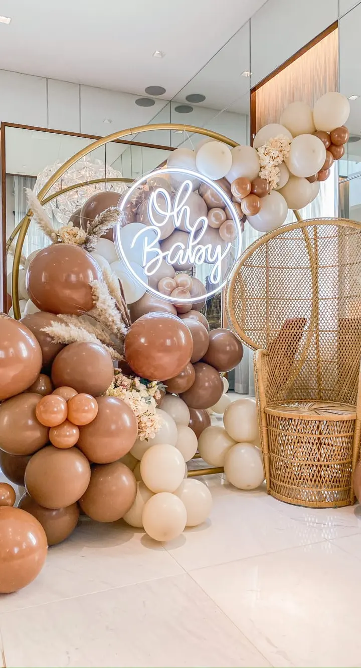 A lavish baby shower display featuring a neon sign that says "Oh Baby", surrounded by an abundance of balloons in various shades of brown and beige, along with a wicker chair and other decorative elements.