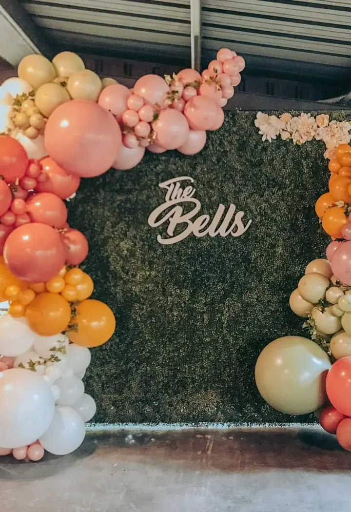 A vibrant and colorful balloon arch surrounds a large black backdrop with the text "The Bells" prominently displayed, creating a visually striking and festive scene.