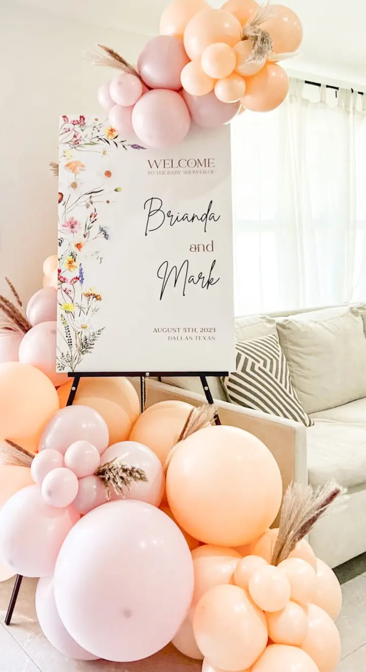 The image depicts a welcoming scene with a large floral arrangement of balloons and flowers in soft, pastel colors, set against a cozy living room backdrop with a sofa and curtains.