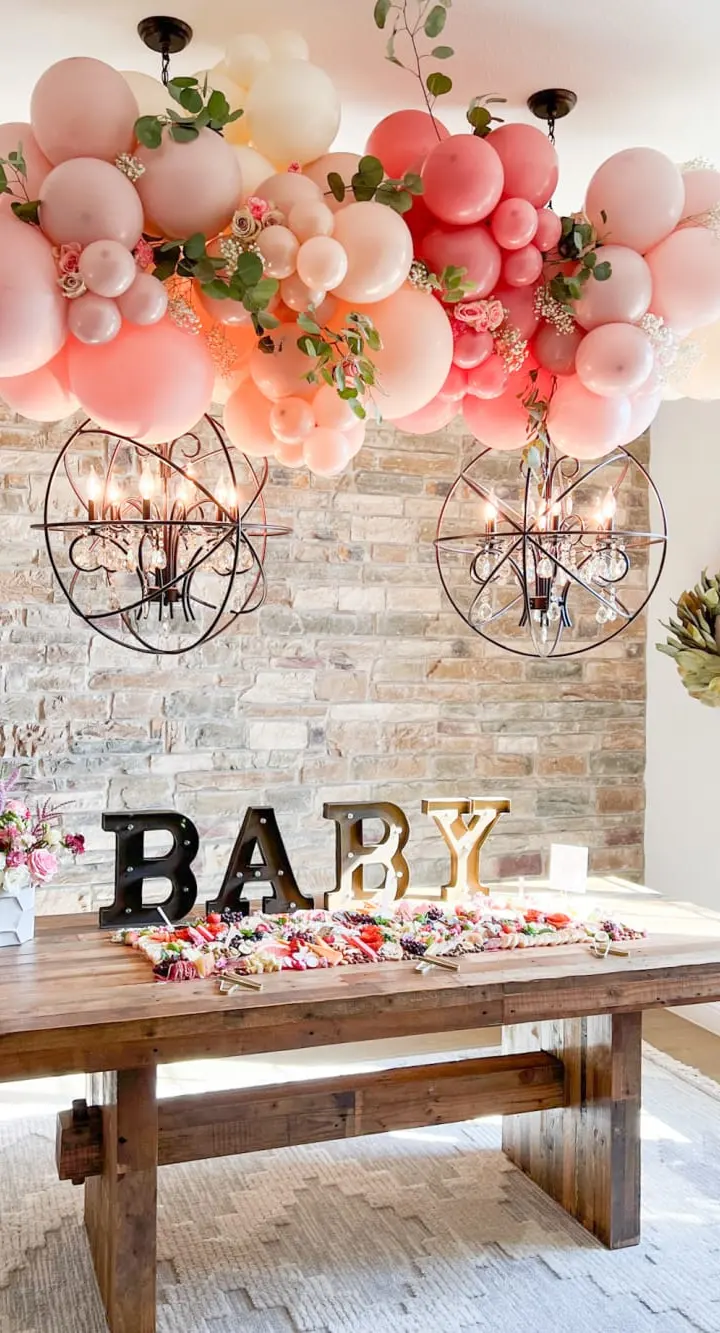 A rustic wooden table adorned with a variety of pink and white balloons, surrounded by a brick wall and complemented by two industrial-style light fixtures, creating a warm and festive atmosphere.