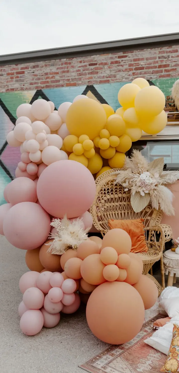 A vibrant display of colorful balloons and decorative elements in the foreground, set against a backdrop of a brick wall with a mural.