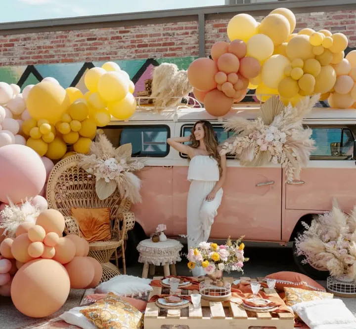 A colorful and whimsical scene featuring a vintage pink van adorned with an abundance of yellow, pink, and white balloons, surrounded by various decorative elements and a festive display.