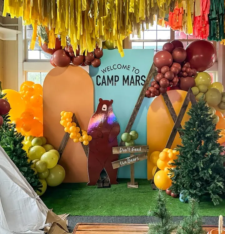 A colorful and whimsical camp-themed display with various decorative elements, including hanging streamers, balloons, and a large welcome sign featuring a bear character, set against a backdrop of lush greenery and a window.
