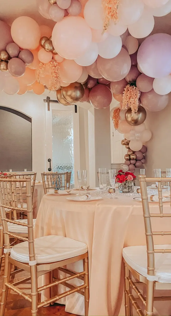 A festive event space with a table set for a celebration, adorned with a vibrant balloon arch in shades of pink, peach, and gold, creating a warm and inviting atmosphere.
