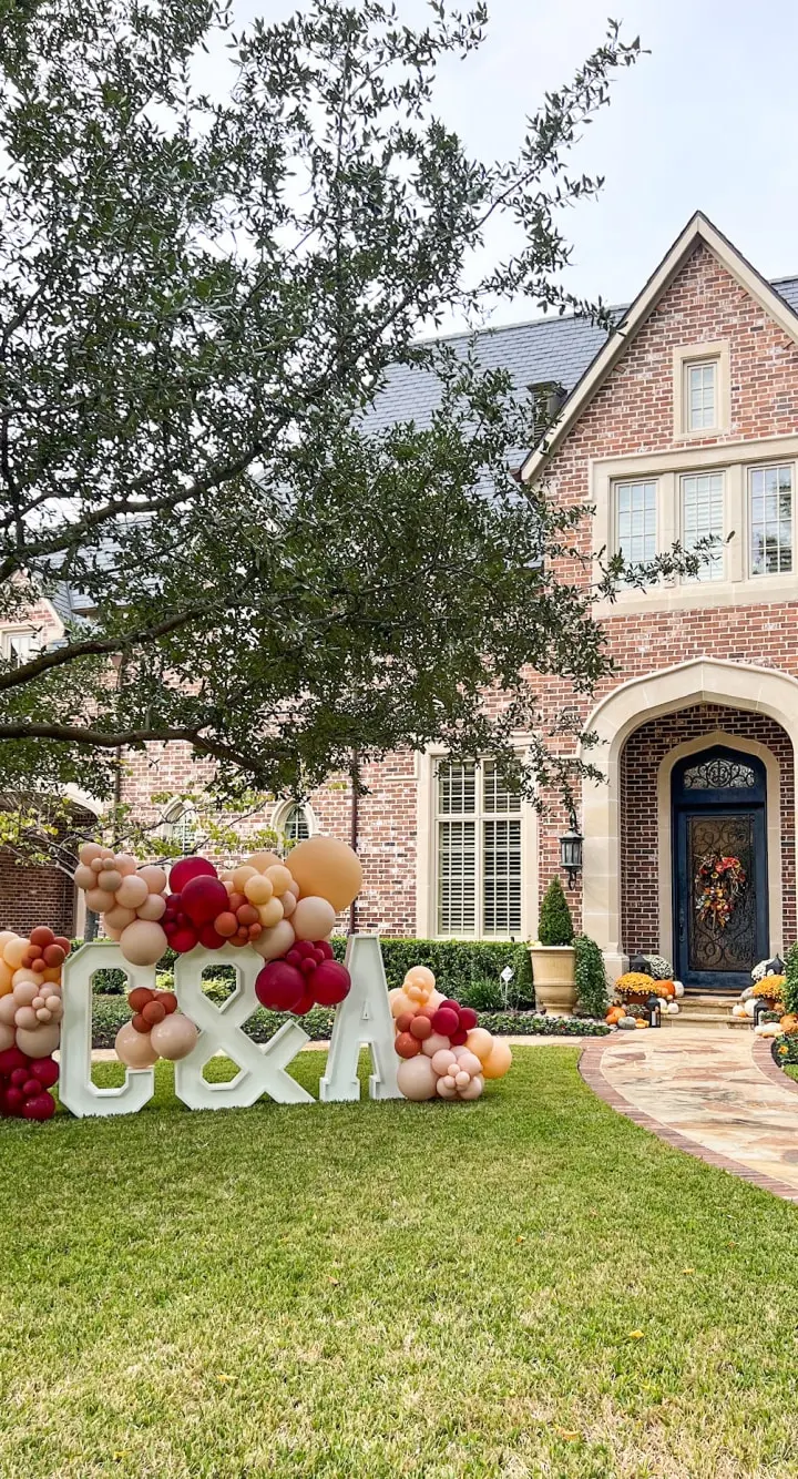 A large, brick house with a decorative entryway is surrounded by a well-manicured lawn, featuring a colorful display of oversized balloons and the word "Bday" in the foreground.