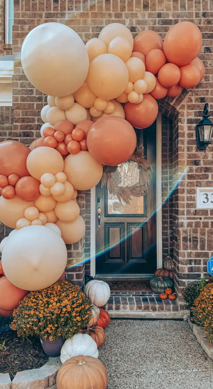 A vibrant display of orange and white balloons adorns the entrance of a brick building, complemented by potted plants and pumpkins on the porch, creating a festive autumn atmosphere.
