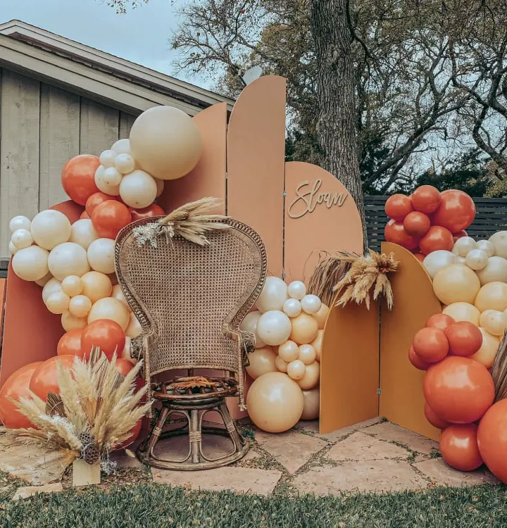 A rustic, autumn-themed display featuring an assortment of decorative elements, including balloons, pumpkins, and woven baskets, set against a backdrop of a wooden structure and lush foliage.