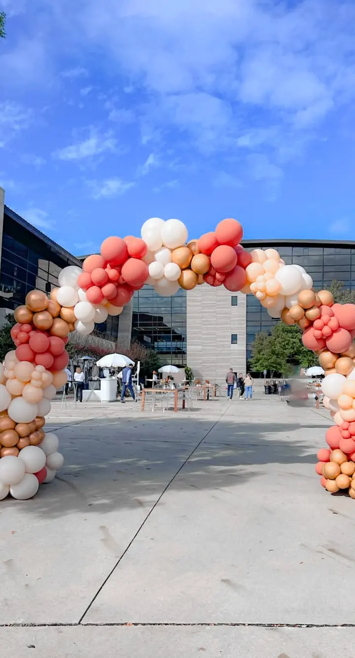 A large, colorful balloon arch stands in the foreground, framing the entrance to a modern, glass-walled building with a blue sky and scattered clouds in the background.