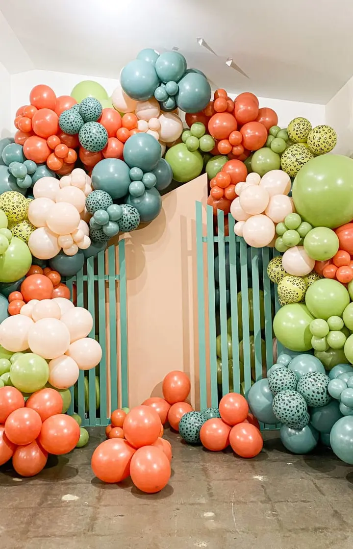 A vibrant and colorful balloon arch made up of various shades of green, orange, and blue balloons, creating a visually striking and festive display in a minimalist indoor setting.