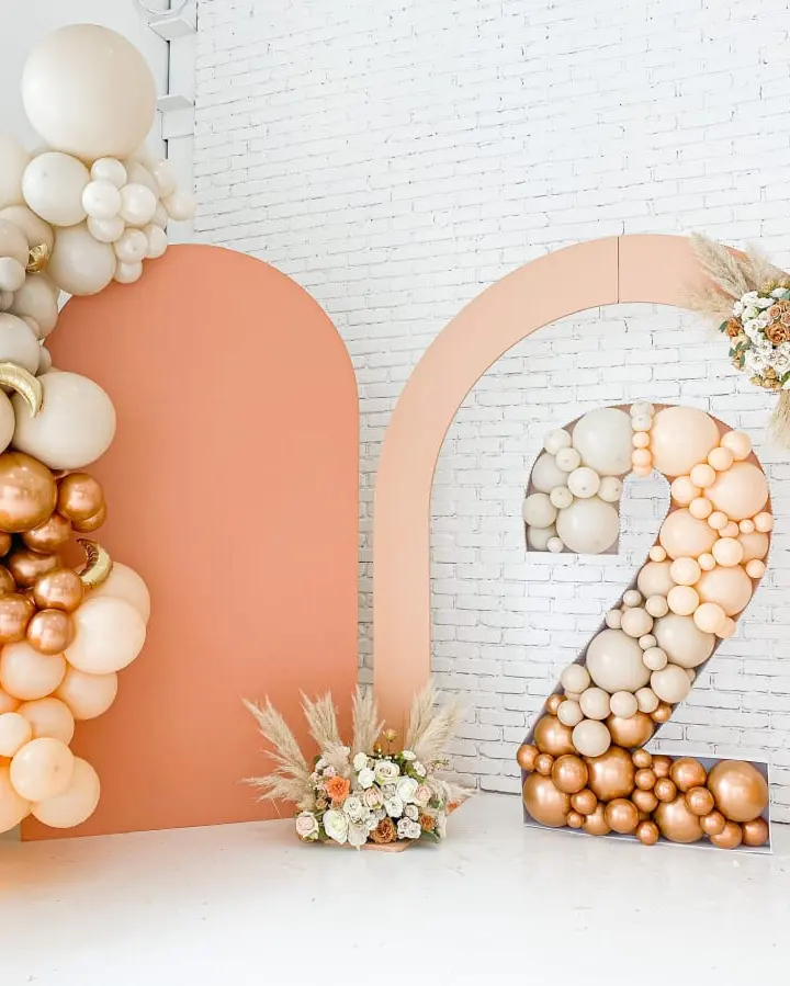 The image features a decorative arrangement of balloons and floral elements in shades of peach, orange, and white, creating a visually appealing and festive display against a plain white brick wall backdrop.
