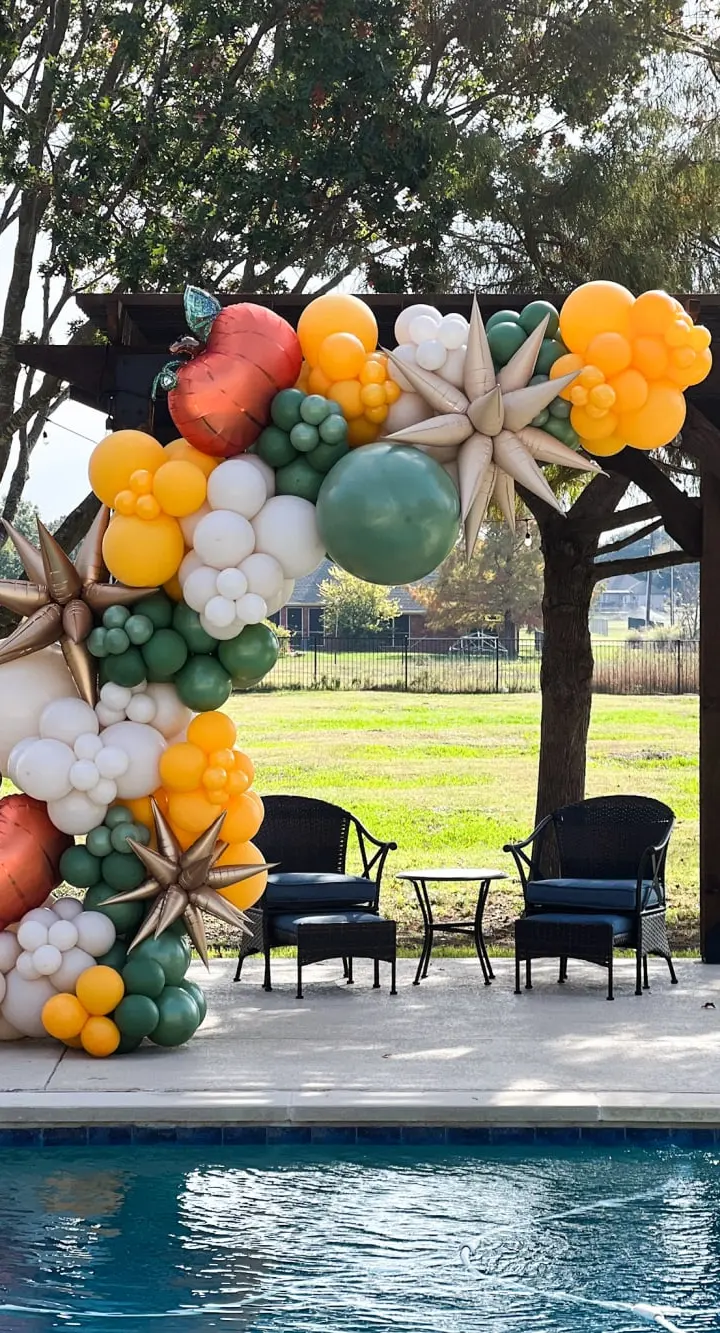A vibrant and whimsical balloon arch adorns the outdoor space, featuring a variety of colorful shapes and sizes, creating a festive and inviting atmosphere. In the background, a lush green lawn and a swimming pool can be seen, adding to the serene and relaxing ambiance of the scene.