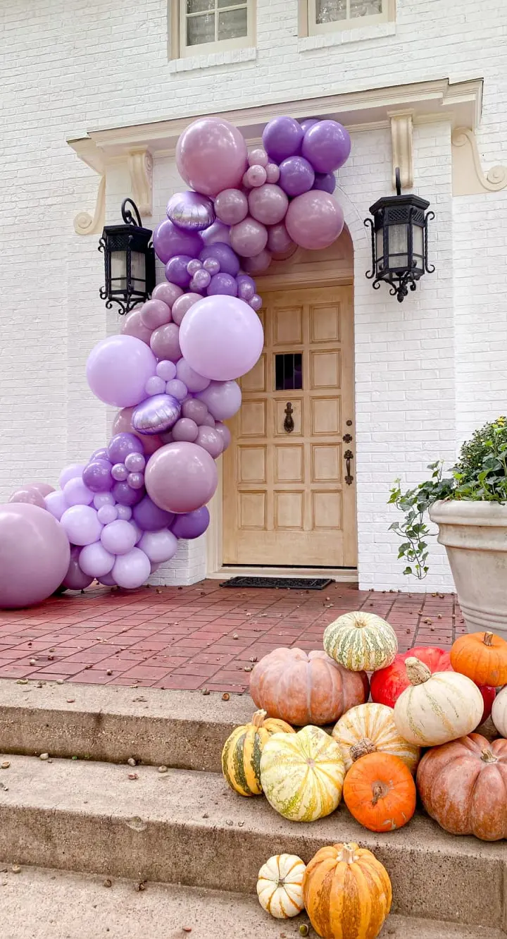 The image shows a grand entrance to a building, adorned with a vibrant display of purple and pink balloons cascading down the steps, complemented by an assortment of colorful pumpkins and gourds arranged in the foreground.