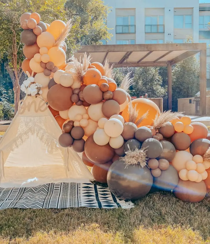 A vibrant and whimsical display of balloons in various shades of orange, brown, and white, set against a backdrop of a building and lush greenery.